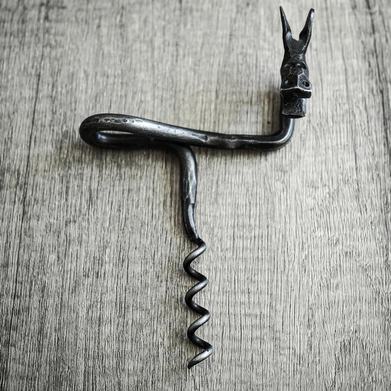 Älgskruv fotograferad ovanifrån på träunderlag. Älghuvudet visar spreade horn, detaljer i metallhantverk synliga. Rektangulär handtag med ögon-detaljer prominent. Antikbörstad stålfinish på mörkmetall mot enkel träbakgrund.