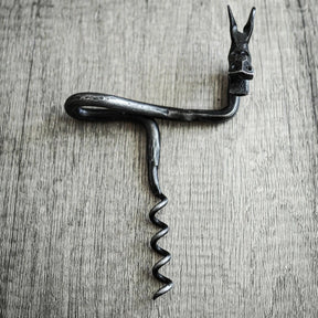 Älgskruv fotograferad ovanifrån på träunderlag. Älghuvudet visar spreade horn, detaljer i metallhantverk synliga. Rektangulär handtag med ögon-detaljer prominent. Antikbörstad stålfinish på mörkmetall mot enkel träbakgrund.