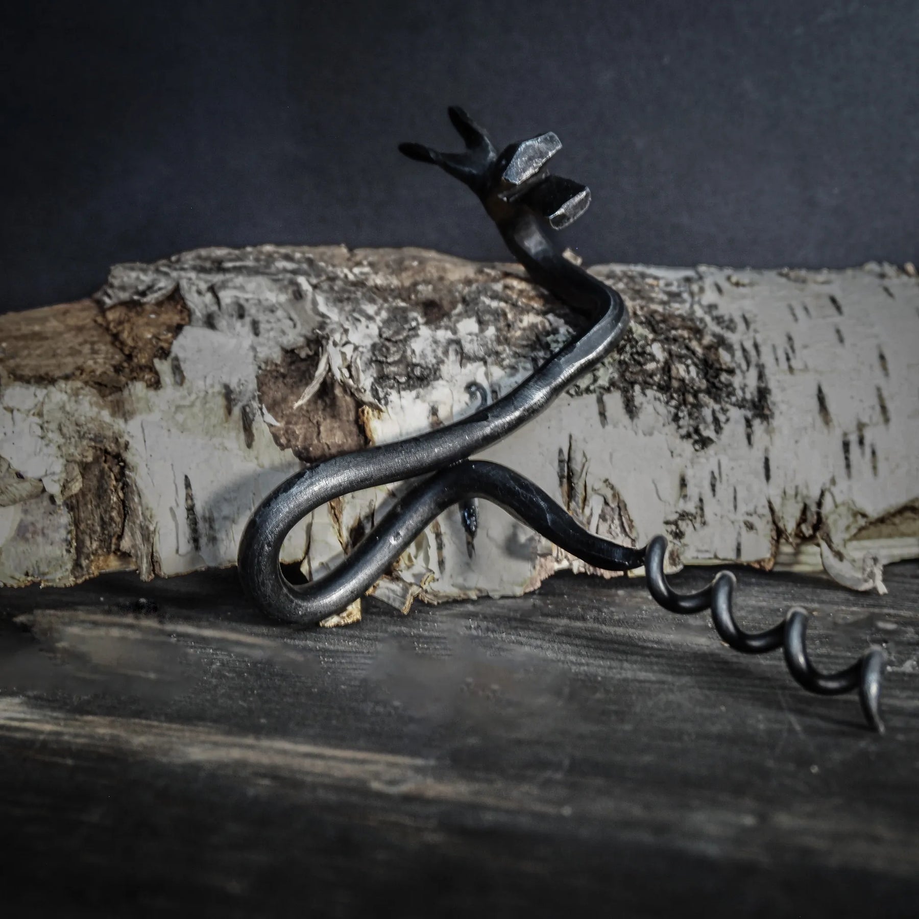 Detaljfoto av handsmide älgskruv. Älghuvudet visar realistiska ögon och detaljer i antikfinish. Metallhandtaget kurvar från älgskulpturen. Fotograferad mot mörkgrå bakgrund med dramatisk belysning som betonar smidets detaljer och textur.