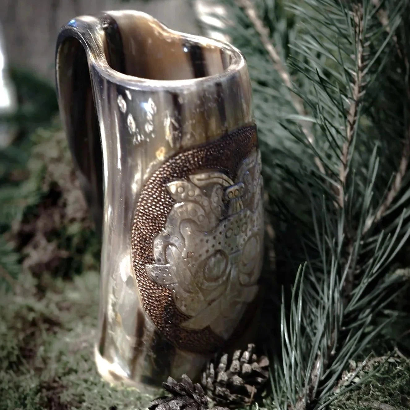 Helkroppsvy av Lokemask hornmugg med tydlig cylindrisk form och funktionell handtag. Mässingsdetaljer på främre ytan visar skulpterad ansikte med fina punktdetaljer. Grön växtlig bakgrund framhäver naturlig estetik.