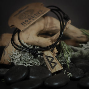 Handgjord runhalsband i ben på svart lädersnöre med Berkano-runan (ᛒ), visad på mannekäng med NORDLYS märke, stiliserad med dried moss, stenar och växter.