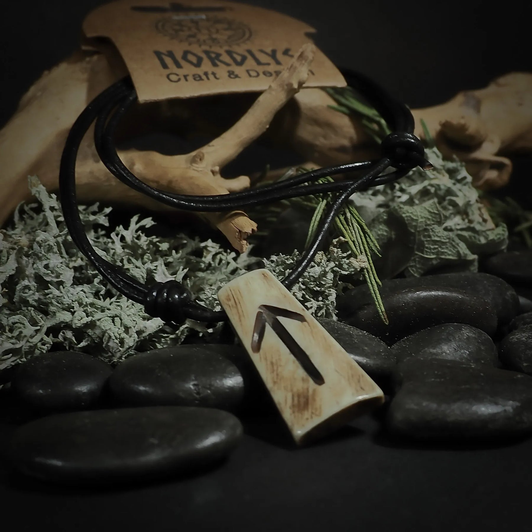Handgjord runornament i ben på svart lädersnodd, visar Fehu-runan från äldfuthark. Fotograferad med svarta stenar och lichen mot mörk bakgrund med varumärkeslapp från Nordlys Craft & Design.