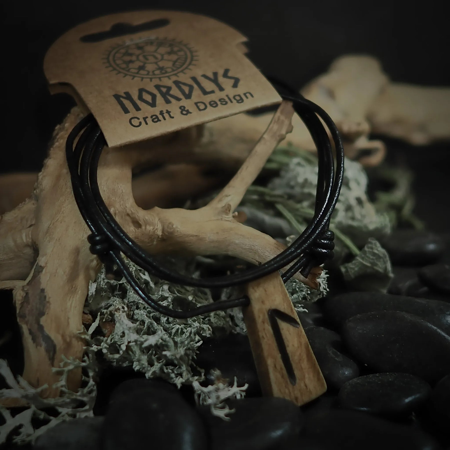 Handgjord runhalsband i ben på svart lädersnöre. Hänget visar en runasymbol inristad i crèmefärgat oxben. Produkten är styladmed mossa och sten på rustik bakgrund. NORDLYS Craft & Design-etikett synlig.