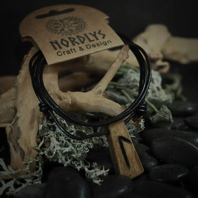 Handgjord runhalsband i ben på svart lädersnöre. Hänget visar en runasymbol inristad i crèmefärgat oxben. Produkten är styladmed mossa och sten på rustik bakgrund. NORDLYS Craft & Design-etikett synlig.