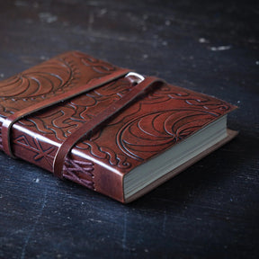 Leather notebook spine with detailed embossed Viking serpent and Triglav-inspired dragon designs, viewed at angle with binding straps visible