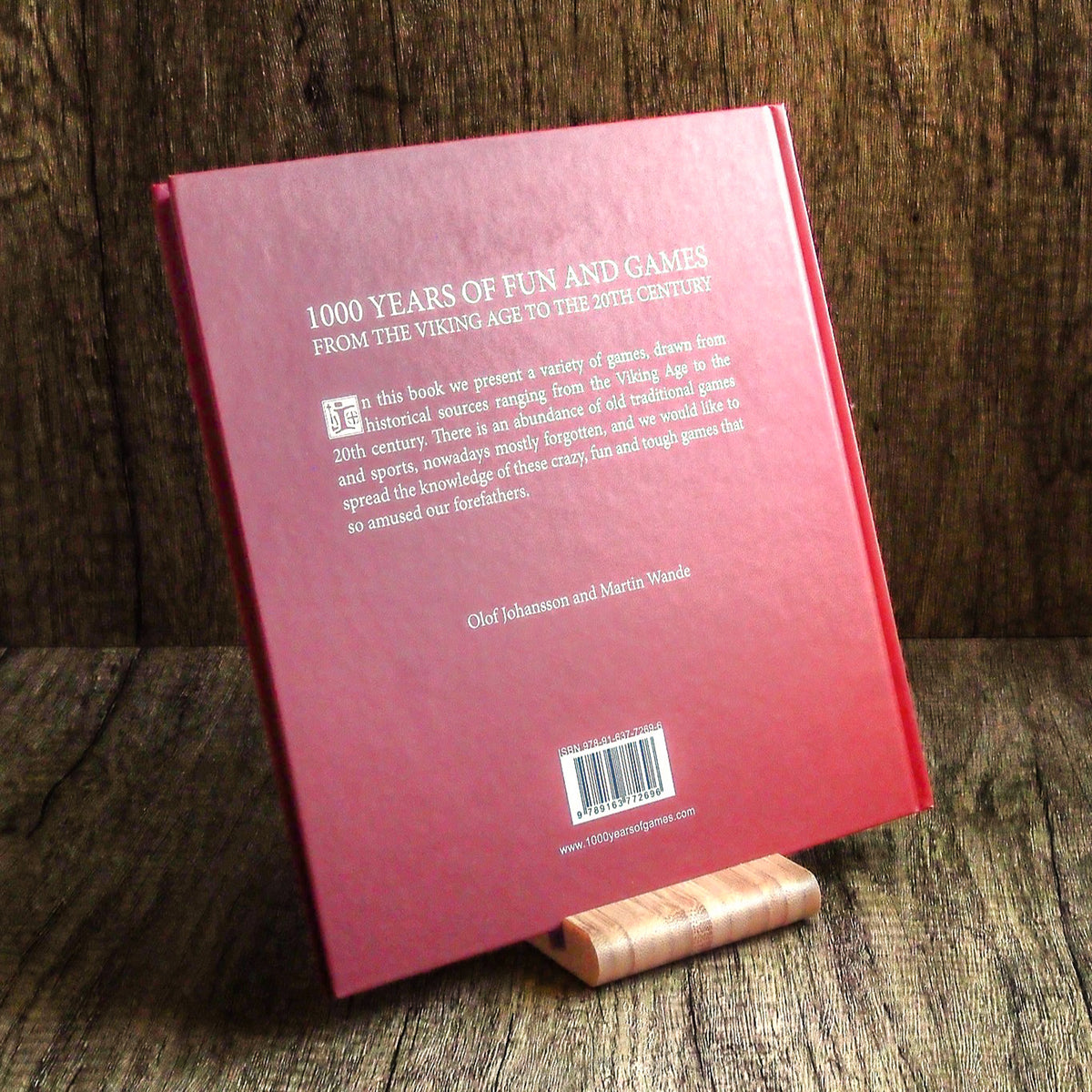 Rosa inbunden bok 'Bok med historiska lekar - 1000 år av nöje och spel' liggandes på trä, visar bakdel med engelsk titel och streckkod