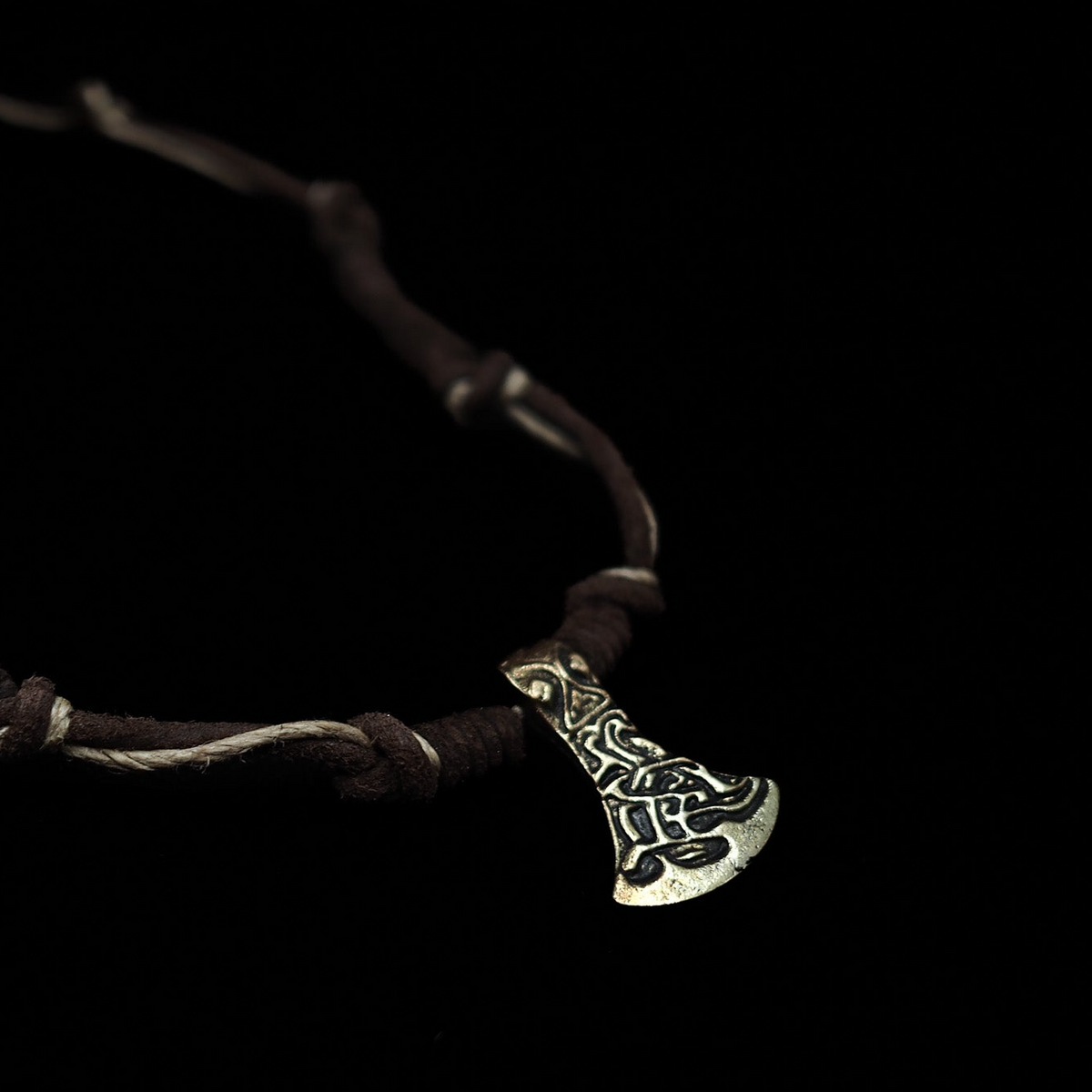 Närbild av ett bronsöxhänge med mycket detaljerade skurkningar och mönster i vikingatida stil, upphängd på ett mörkbrunt läderband mot svart bakgrund.