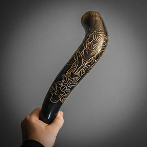 En organisk hornbägare (bock) med mörkbrunt horn och gyllene mässinginlägg. Handgreppet visar graverad bockskalle med noggrant utfört norrskt knotwork och interlacing-mönster. Fotograferad i naturlig miljö med grönska i bakgrunden.