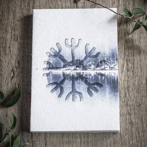 A6 målarbok, Skyddsruna. Öppen uppslag visar Aegishjalmur-rune med speglad skogsmotiv. Fotografi på trä med växter.