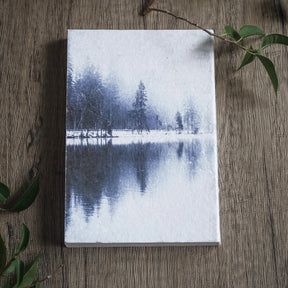 Målarbok A6 med Skyddsruna Aegishjalmur. Sidan visar en lugn vinterscen med snötäckta barrskog och spegeling i vatten, ritad i blå toner på träbakgrund omgiven av gröna växtdelar.