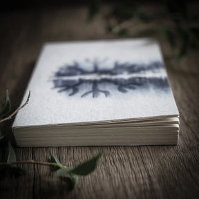 Närbild av målarbok A6-format med Skyddsruna Aegishjalmur. Sidan visar blå barrskog och grangrenar illustrerad på högkvalitativt akvarellpapper, fotad i naturljus.