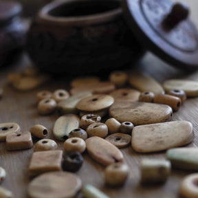 Blandning av vita benpärlor på trä. Pärlorna visar naturlig benåldrande med varierad färgtoning från cremevit till mörkare exponerad benyta. Formerna inkluderar rör, cylindrar, sfärer och rektangulära rummares.