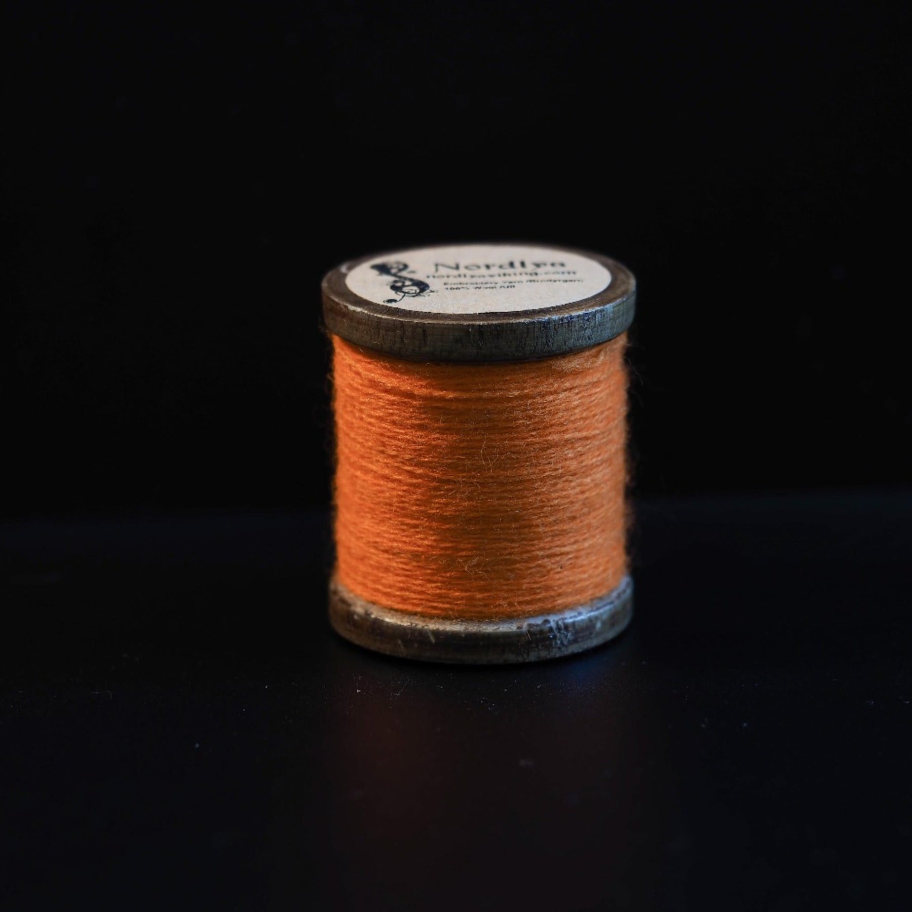 Brændt orange broderitråd på traditionelle trætromler med Nordlys-mærkning. Naturlig jordfarvet tone inspireret af historiske vikingetekstilfarver.