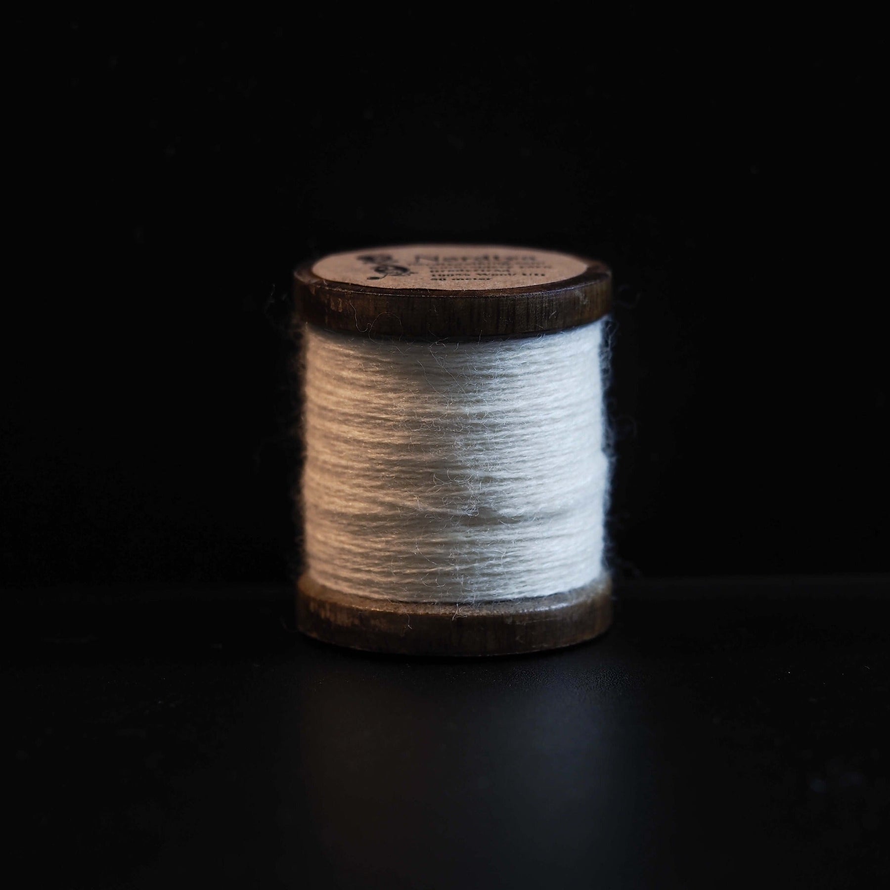 En enkel spole med lysegrå uldgarn på mørk baggrund, viser højkvalitets broderitråd med træspole og etiket