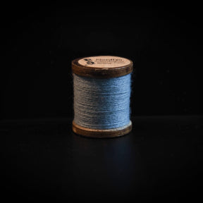 En enkel spole med blågrå uldgarn på mørk baggrund, viser nordisk farvepalet og højkvalitets broderitråd med træspole