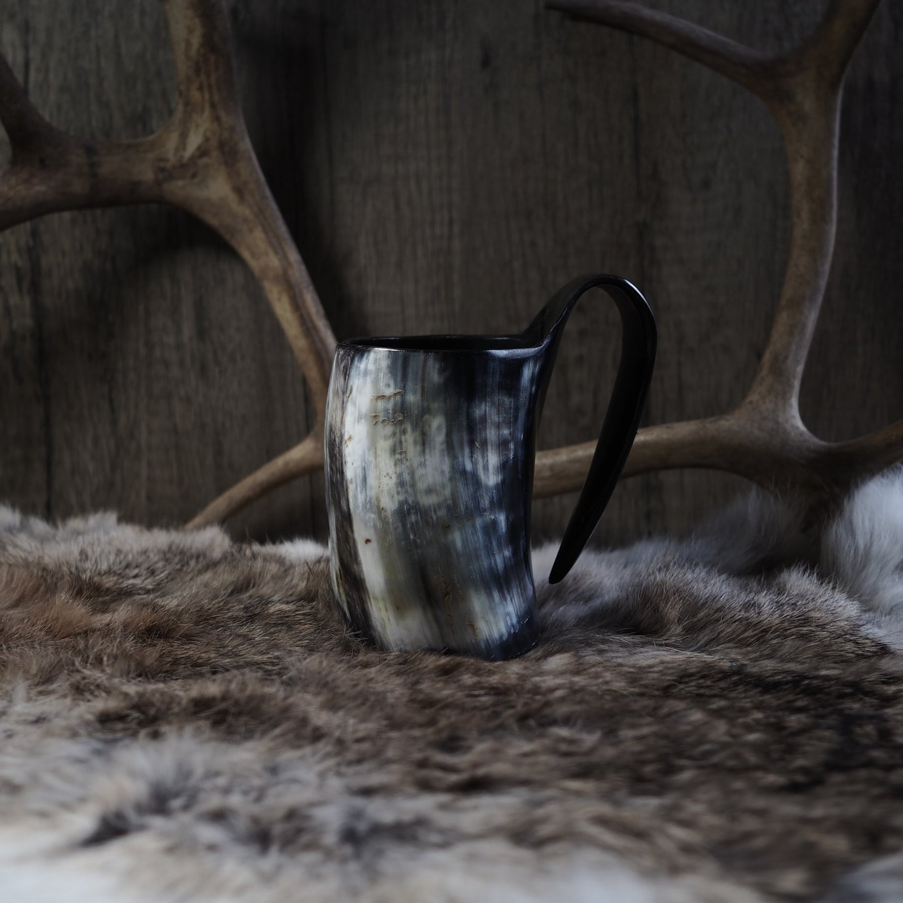 Händer som håller en mörk hornmugg med varm dryck, med läder- och metallarmband synliga. Visar muggens användning i naturlig miljö.