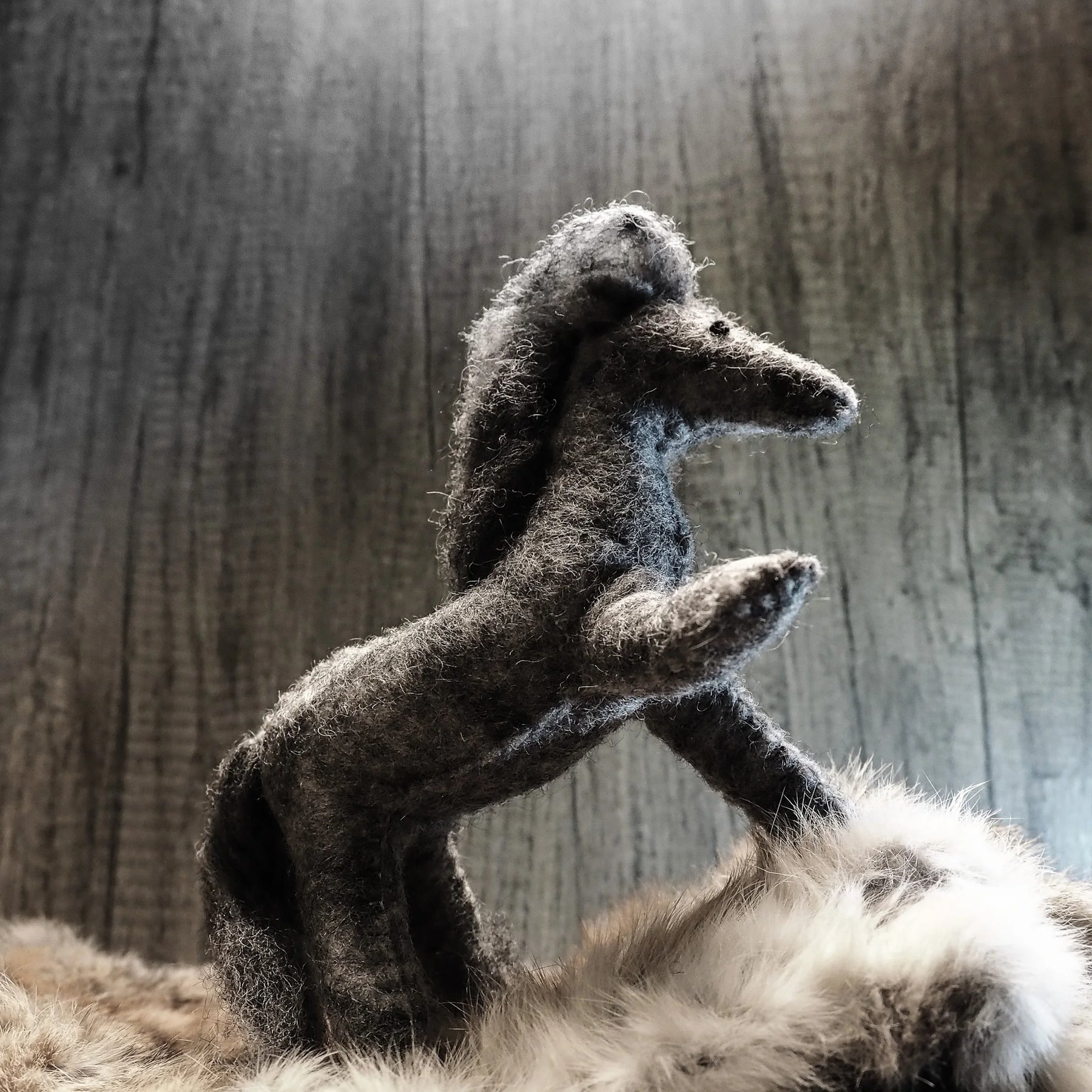 Stor grå hund i ullfiltat hantverk som presenteras med framben upphöjda i dynamisk hållning. Detaljerad ull-textur över hela kroppen med fluffigt man och svans. Bakgrund av slitet grått trä. Trådförstärkning i benen möjliggör posering.