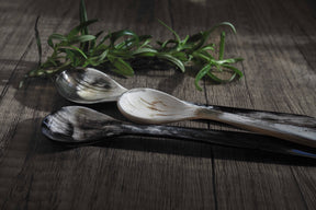 Tre långa teskear i ko-horn med flerfärgade mönster, visar naturliga mörkbruna, kremfärgade och svarta ränder. Presenterade på grå trägolv med färska rostmarvskvistar.