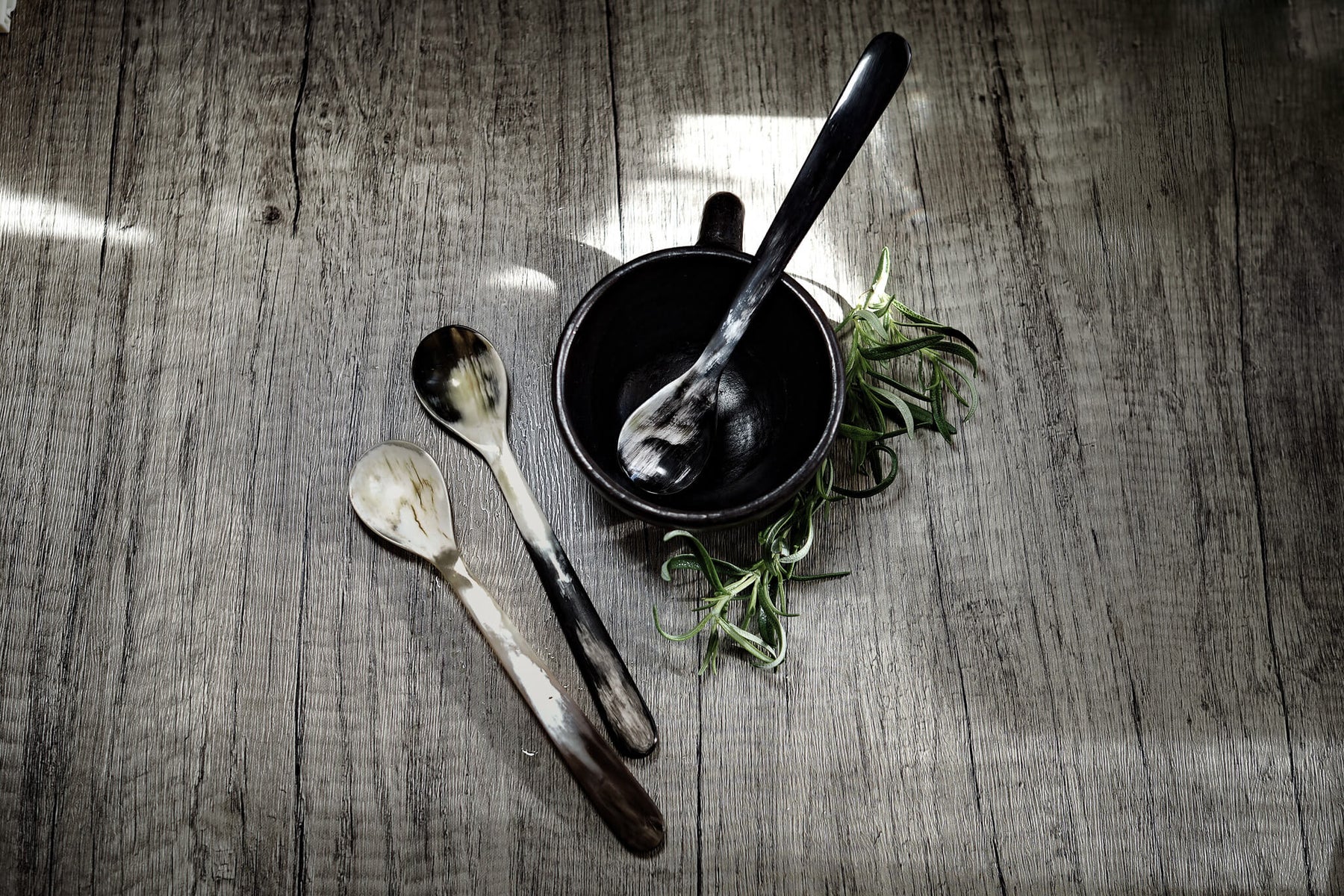 Lång tesked i flerfärgad ko-horn med naturliga mörkbruna och kremfärgade ränder, presenterad i en svart skål med färska rostmarv på grå trägolv.