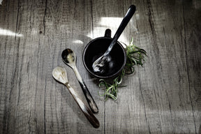 Lång tesked i flerfärgad ko-horn med naturliga mörkbruna och kremfärgade ränder, presenterad i en svart skål med färska rostmarv på grå trägolv.