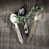 Fyra bestickdelar i horn med naturlig färgvariation och glansiga ytor, arrangerade symmetriskt på trä med grön örthärsmak