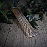Ljus kamm i oxhorn med fina, tätt sittande tänder. Tillverkad av organiskt horn utan statisk laddning, helt nedbrytbar. Visad på rustik grå träyta med gröna växter.