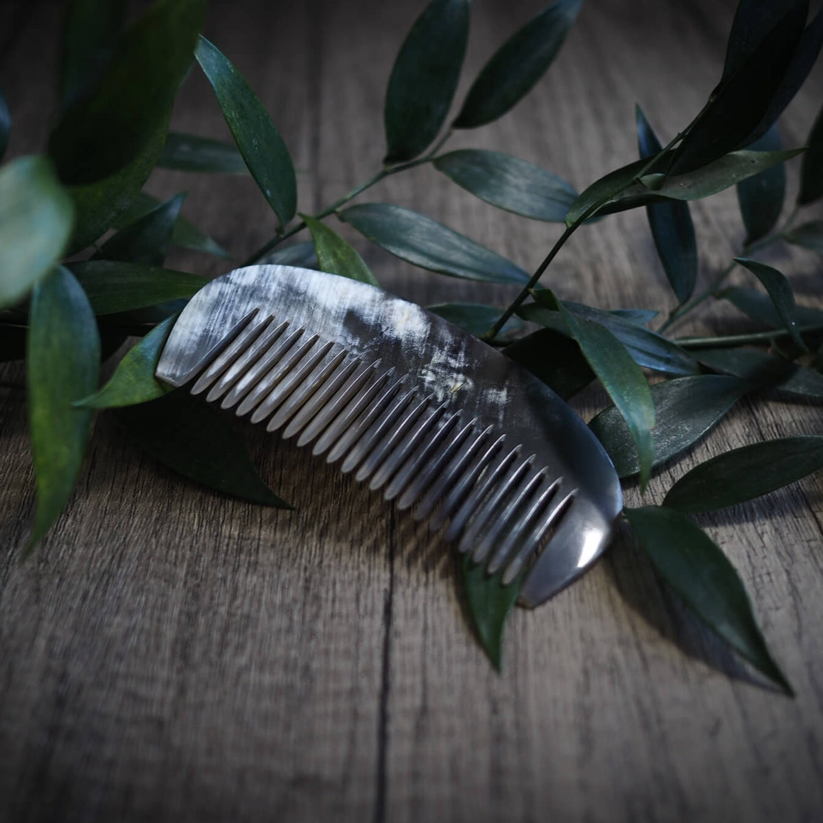En mörk hornsked med finspetsade tänder, omgiven av gröna växtgrenar på en träyta. Produktfoto för vikingisk hårkam av naturligt horn.