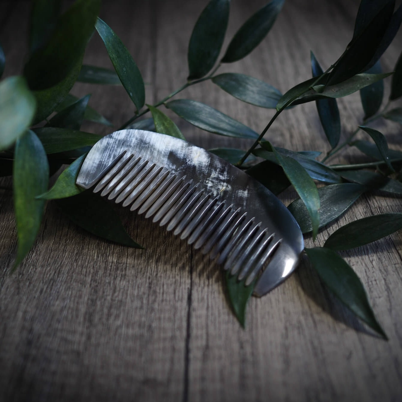En mörk hornsked med finspetsade tänder, omgiven av gröna växtgrenar på en träyta. Produktfoto för vikingisk hårkam av naturligt horn.