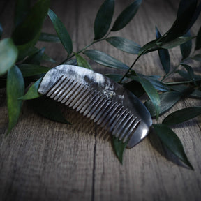 En mörk hornsked med finspetsade tänder, omgiven av gröna växtgrenar på en träyta. Produktfoto för vikingisk hårkam av naturligt horn.