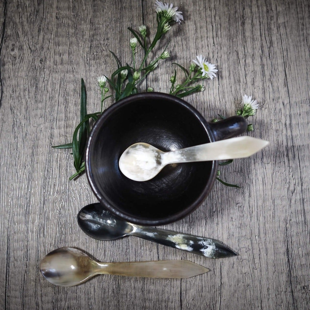 Hornbestick med smörknivar och sked arrangerade med svart keramikskål, vita örter och blomster på grå träyta