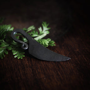 Kvinnokniv mini på mörk snöre placerad på träyta med grönt örtkrynt. Visa miljösamband och naturlig presentation av hängesmycket.