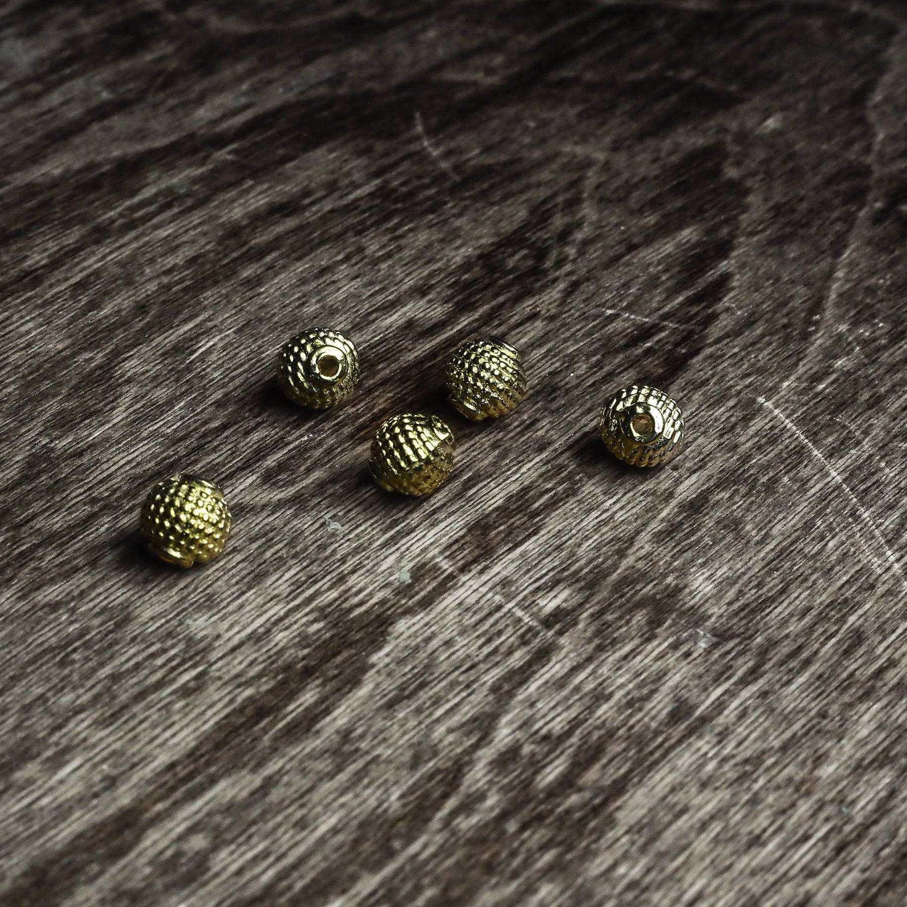 Fem guldfärgade Freja-pärlor med granulerad yta utspridda på mörkt grått trä