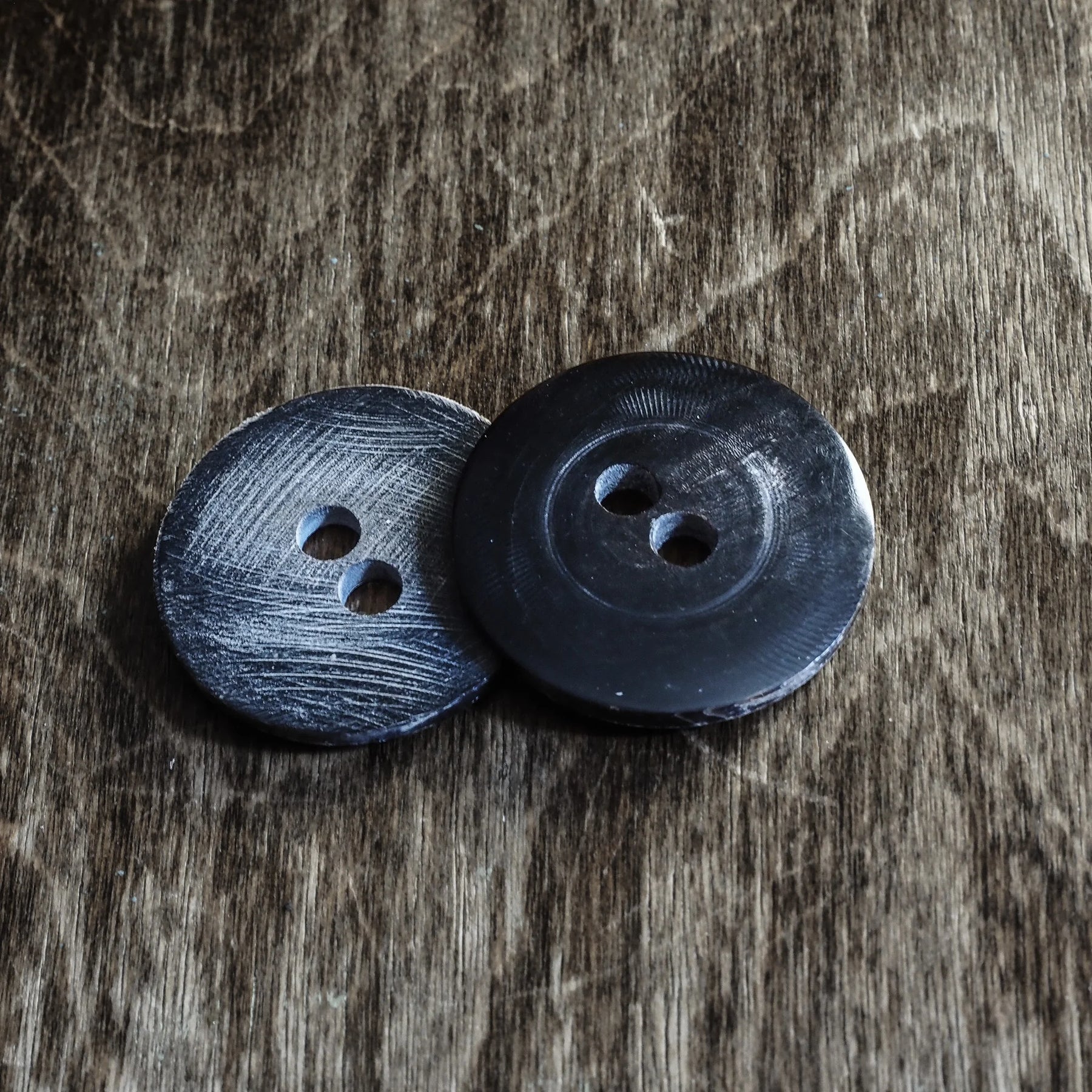 Två hornknappar på slitet grått träunderlag. Vänster knapp visar borstad finish med synlig kornstruktur och två håll. Höger knapp är polerad, mörkare och glänsande med två sänkta håll. Båda cirka 35 mm i diameter och 2 mm tjocka. Organiskt material från hållbar horn.