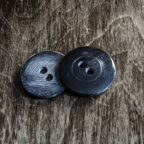 Två stora hornknappar på vintage grått träunderlag. Vänster knapp har borstad finish med synliga kornlinjer. Höger knapp är polerad och glänsande med mörkare färg. Båda knappar har två håll för sy och är cirka 35 mm i diameter. Gjorda av organisk, hållbar horn.