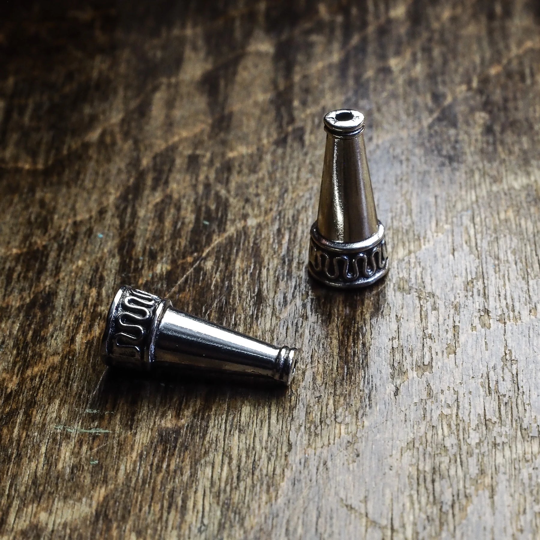 Två silverpläterade ytterpärlor med konisk form på träyta. Visar detaljerad dekorativ ring och mässingsarbete vid basen av varje pärla.