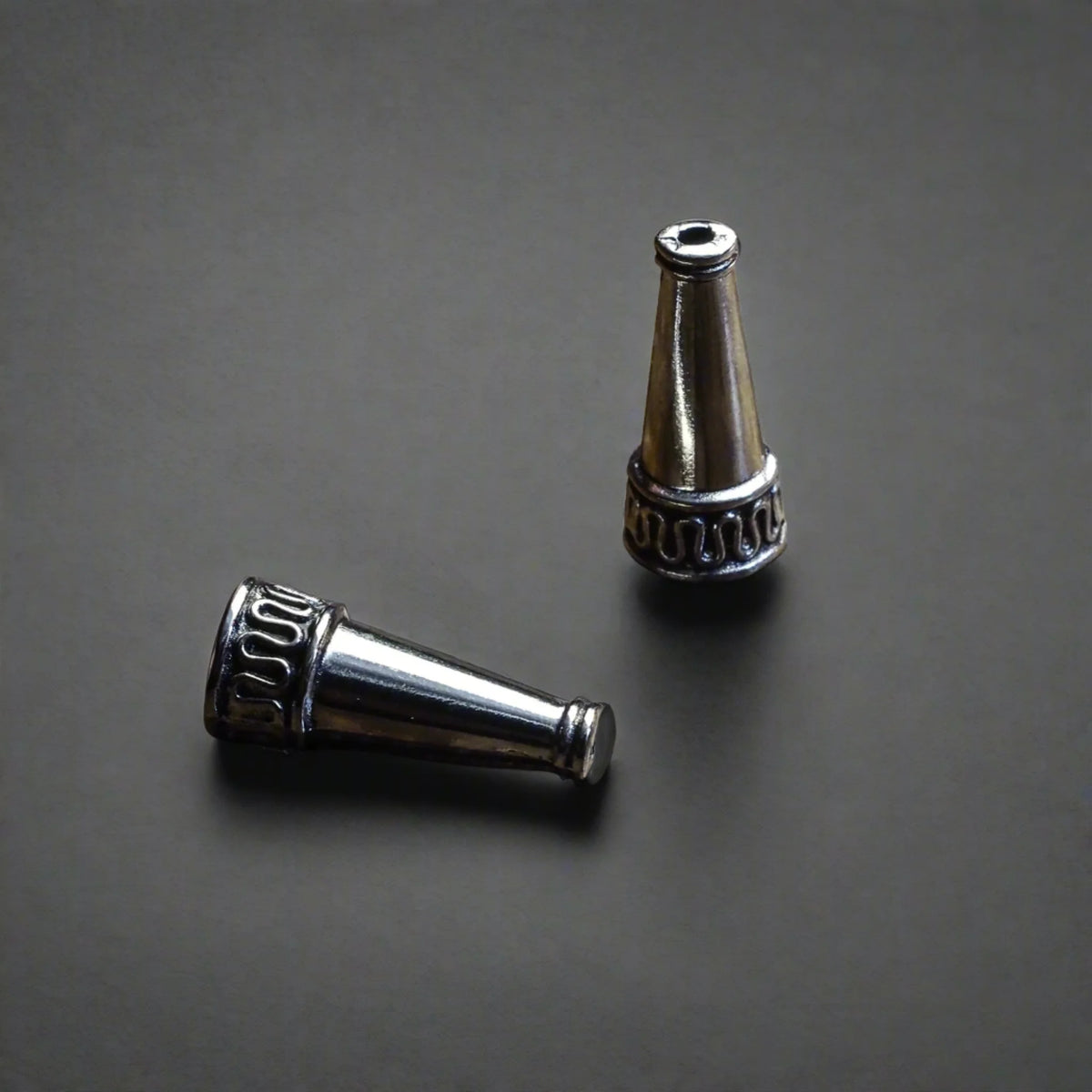 Två silverpläterade ytterpärlor med konisk form, fotograferade från olika vinklar på grå bakgrund. Visar dekorativ ring och detaljerat arbete på mässingsperlan.