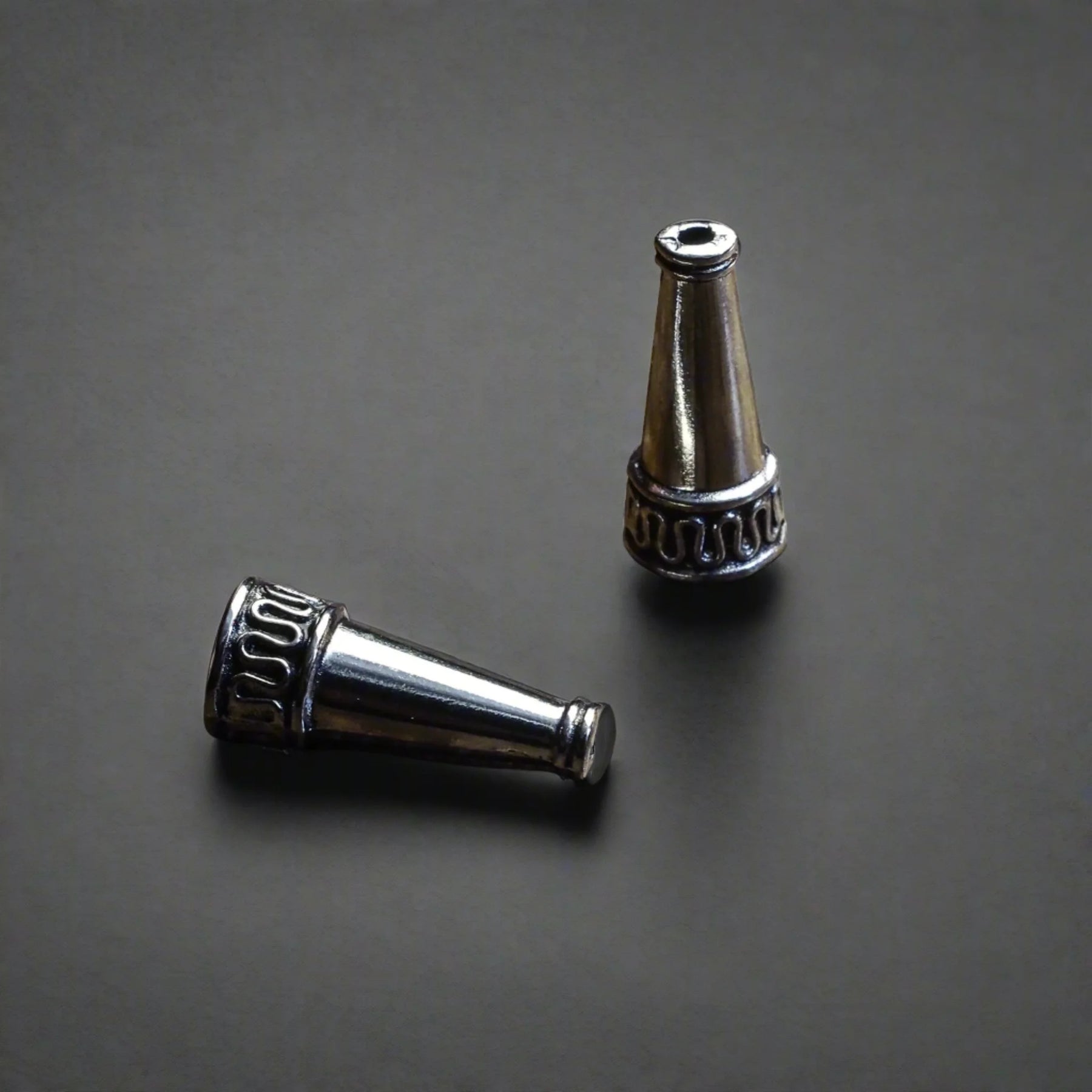 Två silverpläterade ytterpärlor med konisk form, fotograferade från olika vinklar på grå bakgrund. Visar dekorativ ring och detaljerat arbete på mässingsperlan.