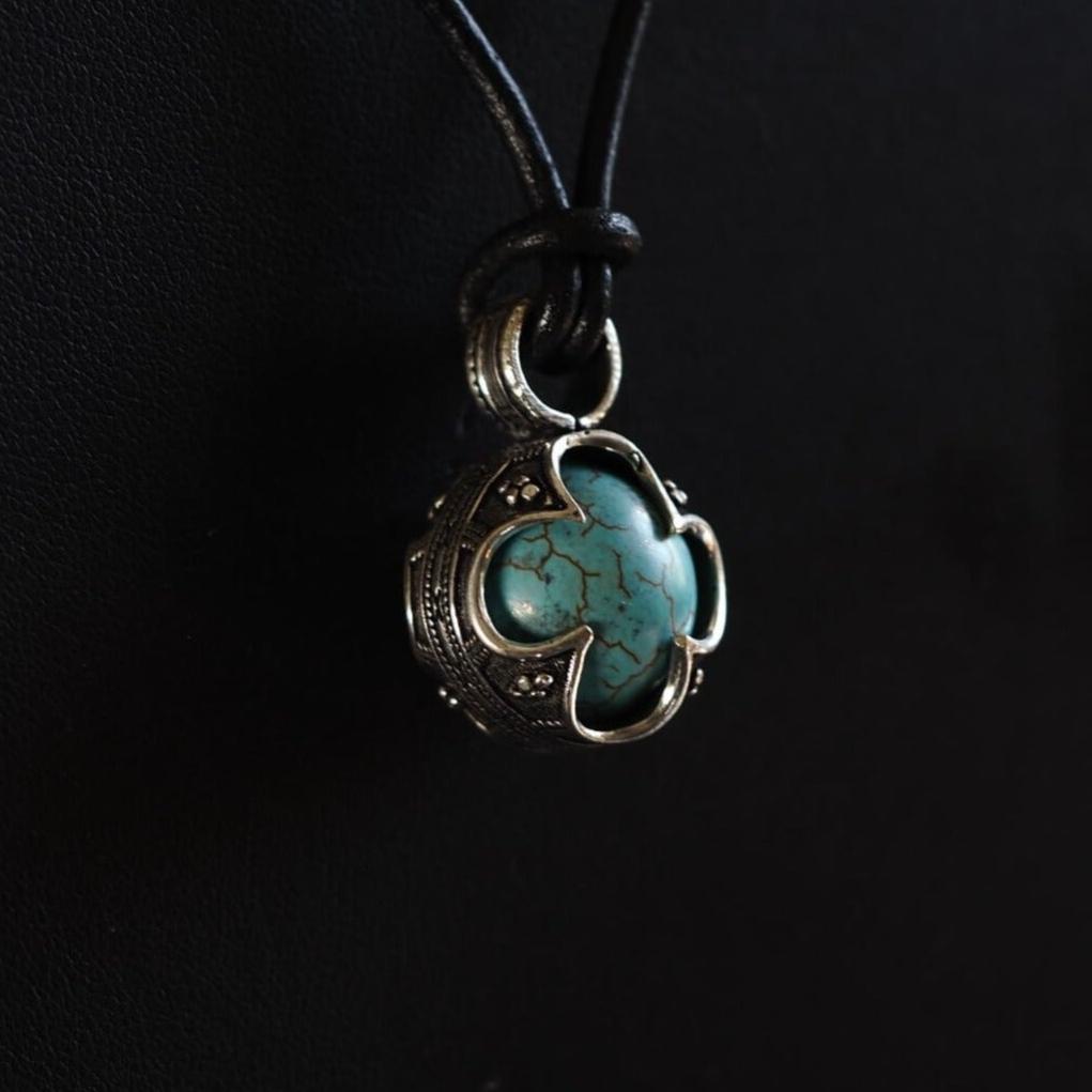 Turkos hängsmycke med graverad mässing och flätat svart snöre. Stenartefakten visar naturliga sprickmönster. Mässingsfattningen är dekorerad med keltisk knutverk och skyddslobber.