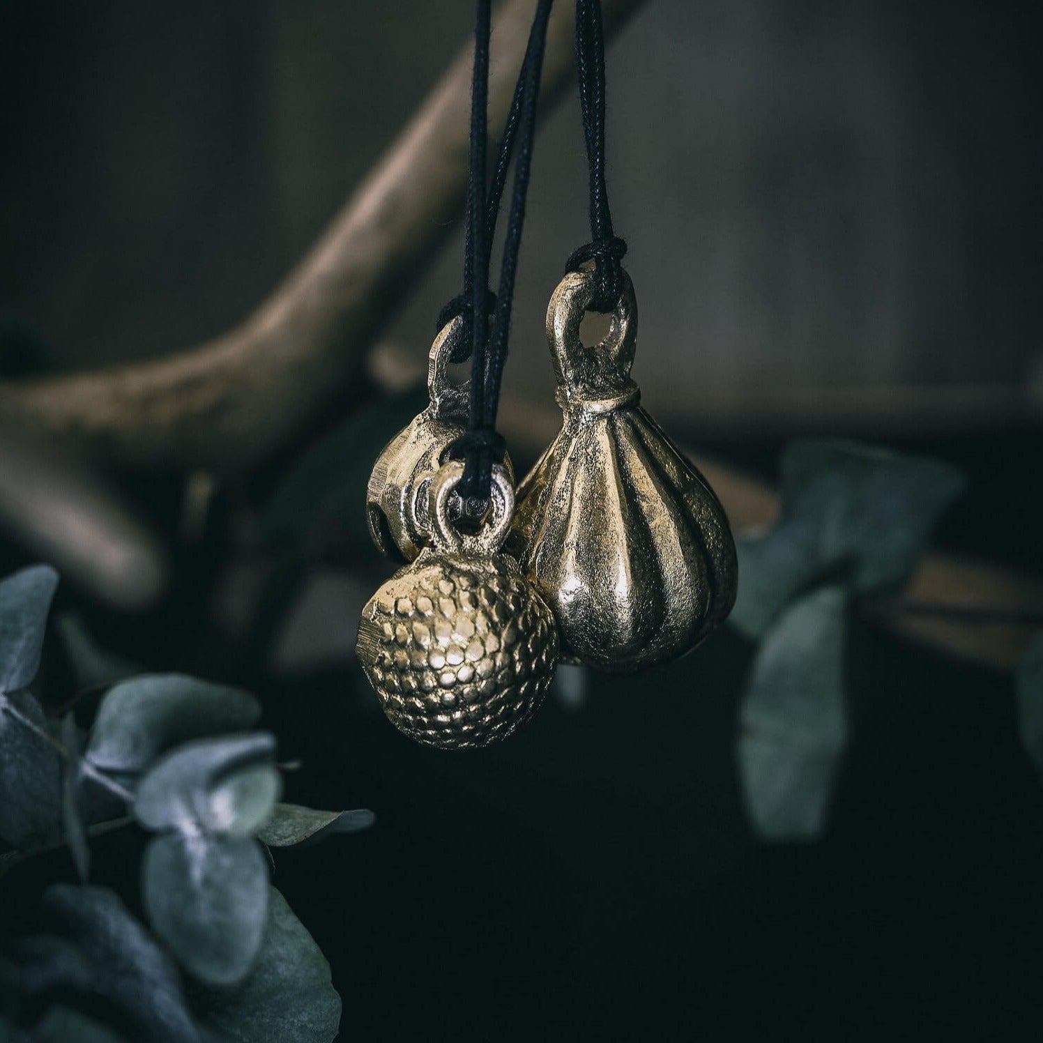 Tre mässingklockor hängande på svart snöre mot naturlig buskbakgrund. Klockorna visar olika mönster: spiraler, släta teardropformer och prickad textur. Varierande designs i samma produktserie.