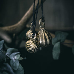 Tre mässingklockor hängande på svart snöre mot naturlig buskbakgrund. Klockorna visar olika mönster: spiraler, släta teardropformer och prickad textur. Varierande designs i samma produktserie.