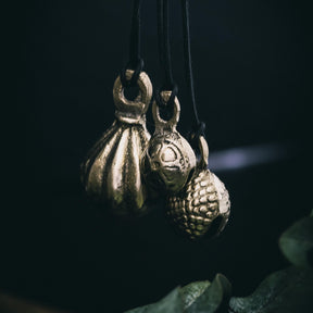 Tre mässingklockor hängande på svarta snören. Varierande ytfinish: radiella ribbor, groplad textur och reliefmönster. Grön naturlig bakgrund. Professionell smyckesstyling med studiebelysning.