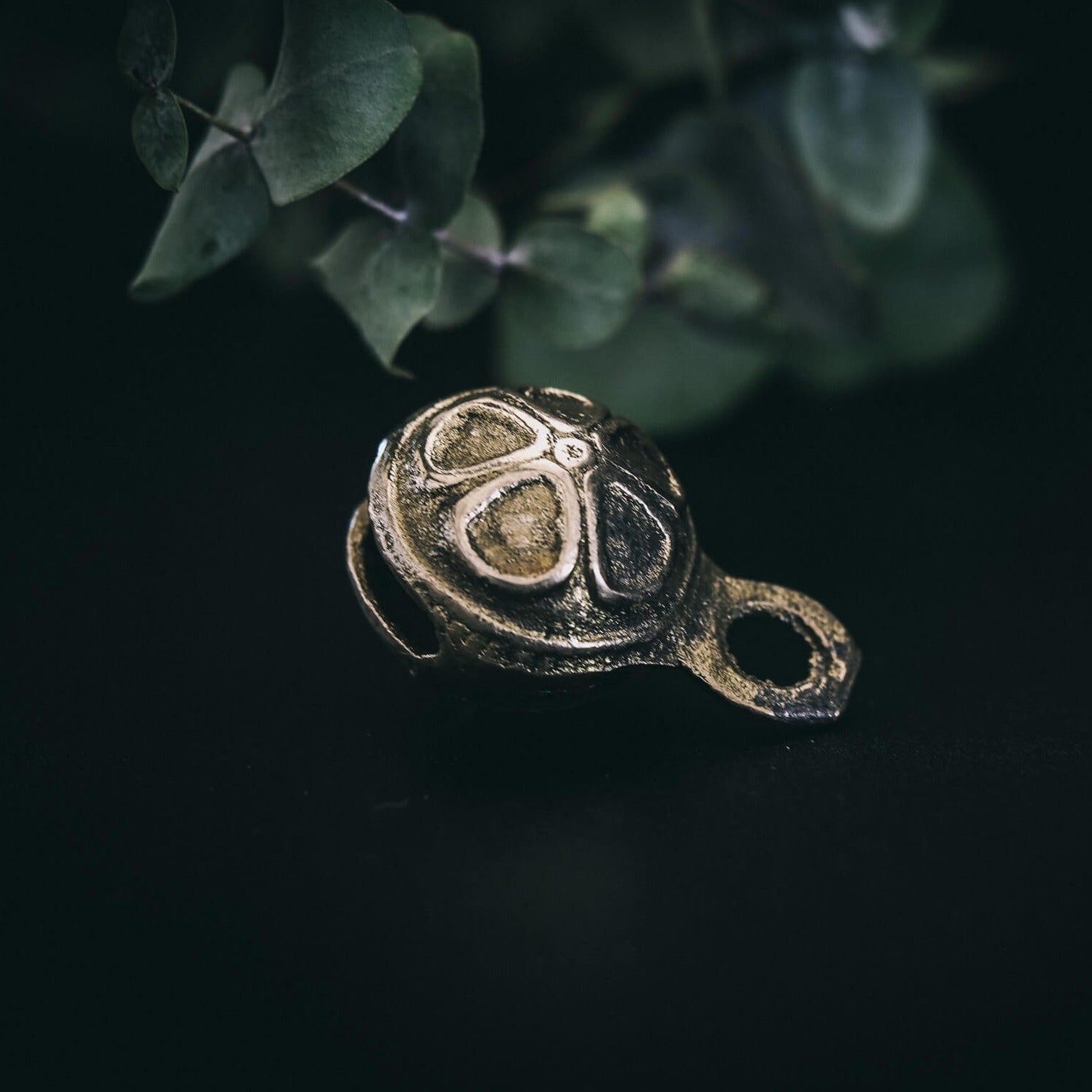 Mässingklocka med 30mm diameter och dekorativ prickad yta på mörk bakgrund, hängande från ögla. Närbildsfotografi som visar metalglans och texturerad design.