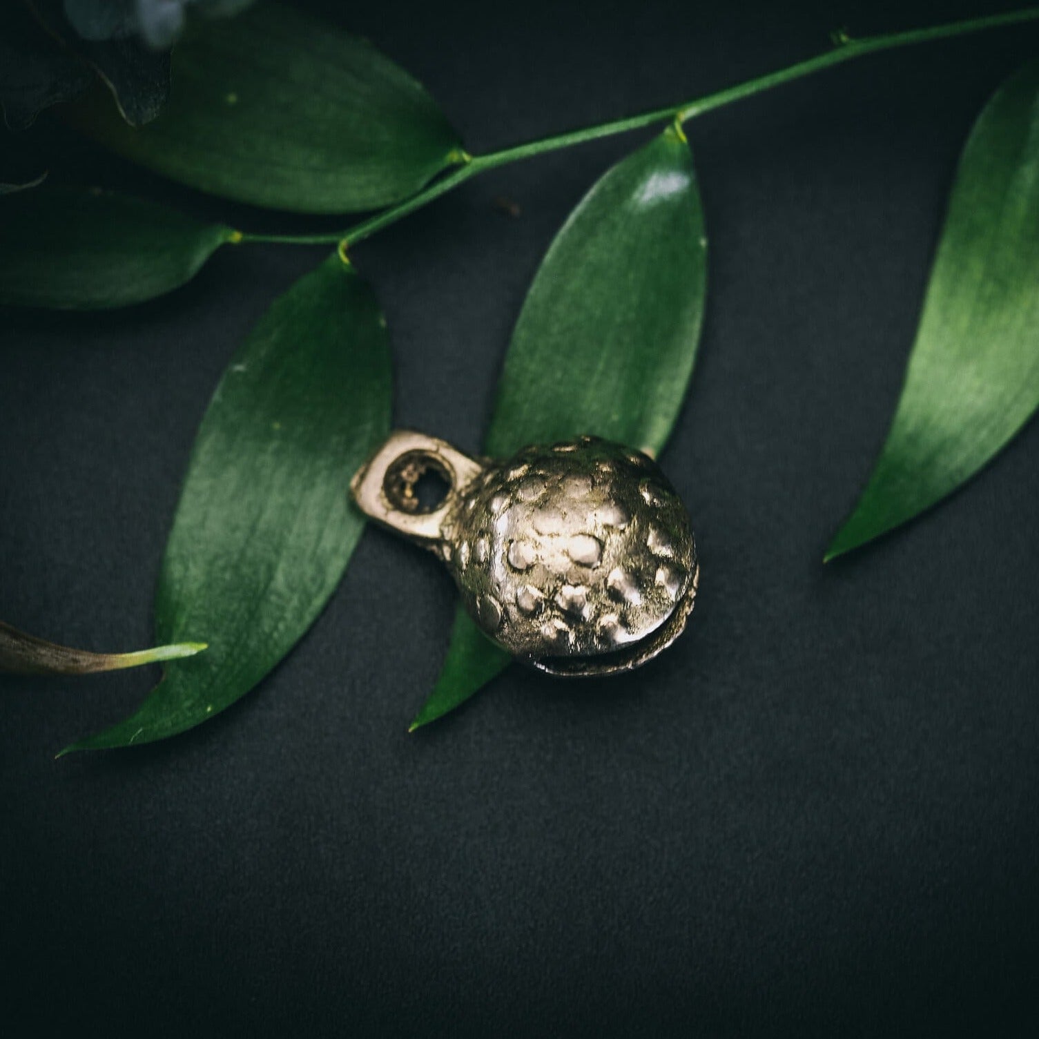 Mässingklocka i närbild med tydlig prickad och groplad yta. Små cirkulära reliefmönster täcker hela ytan. Snöre genom ögla synligt. Naturlig belysning visar metallglans och hantverkskvalitet.