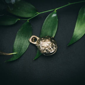 Mässingklocka i närbild med tydlig prickad och groplad yta. Små cirkulära reliefmönster täcker hela ytan. Snöre genom ögla synligt. Naturlig belysning visar metallglans och hantverkskvalitet.