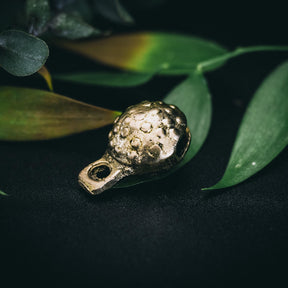 Mässingklocka placerad på gröna växtblad. Klockans yta visar tydlig prickad och groplad textur. Varm metallglans och naturlig belysning. Handgjord skandinavisk stil.