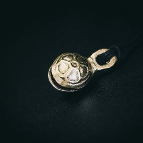 Mässingklocka hängande på svart snöre mot mörk bakgrund. Ytan visar radiella ribbor och groplad textur. Metallglans framhävs av studiabelysning. Ren minimalistisk produktvy.