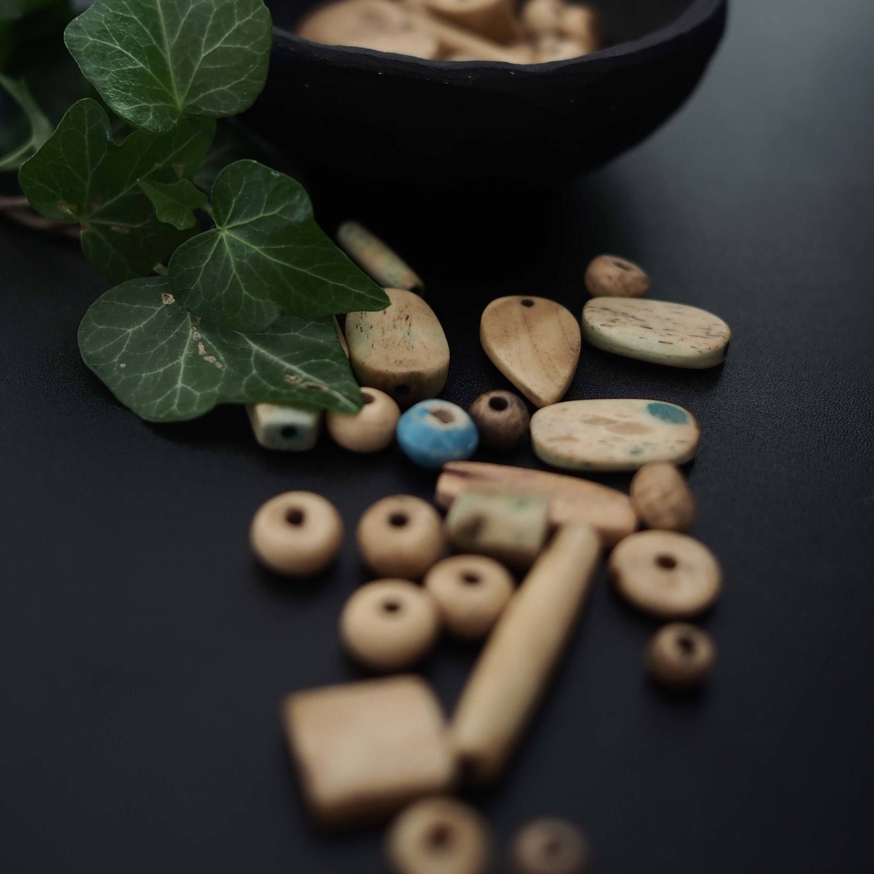 Blandning av vita benpärlor arrangerade på mörk yta med gröna växter. Pärlorna visar naturlig benåldrande och varierar mellan sfärer, oval formade och cylindriska rör med hand-snidtad asymmetrisk karaktär.