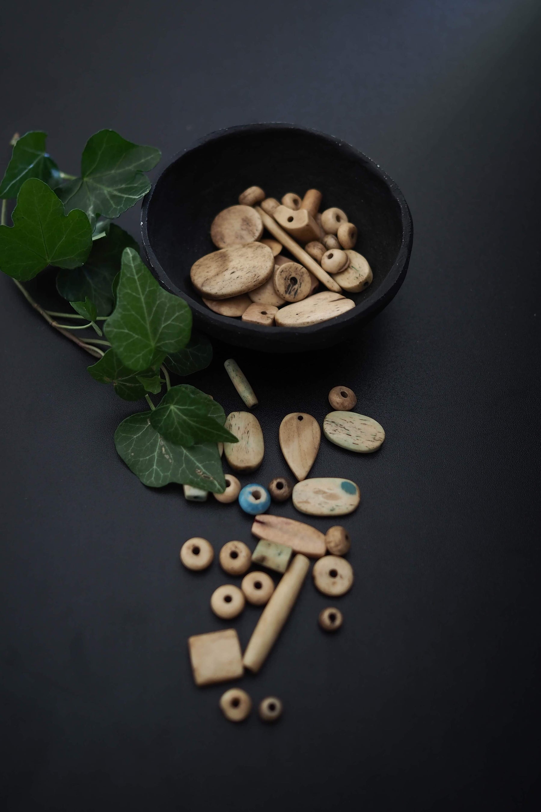 Blandning av vita benpärlor på mörk bakgrund med gröna växter och svart keramikskål. Pärlorna omfattar sfärer, fat-formade rör, cylindrar och rektangulära rummares med naturlig benåldrande och hand-snidad karakteristika synlig.