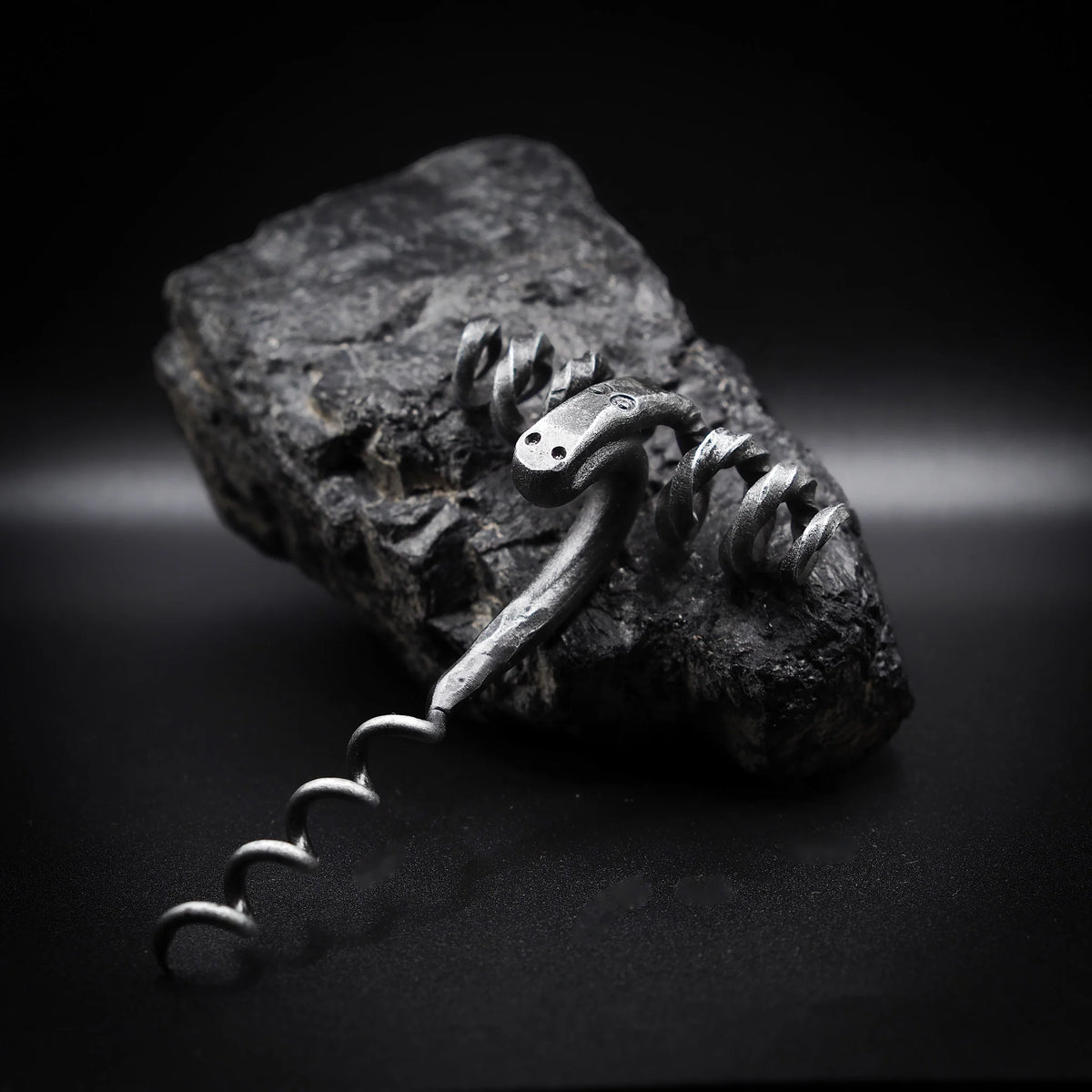 Handsmide bagge-skruv (råmskruv) på granitatsten. Gjord av återvunnet järn med mörkpatina och metalliska högdagrar. Baggehuvudet visar böjda horn som formar dramatisk handtag. Ansiktsfötryck och öga-detaljer synliga. Spiral skruv nedan. Dramatisk sidbelysning betonar metallartverk och hantverk.