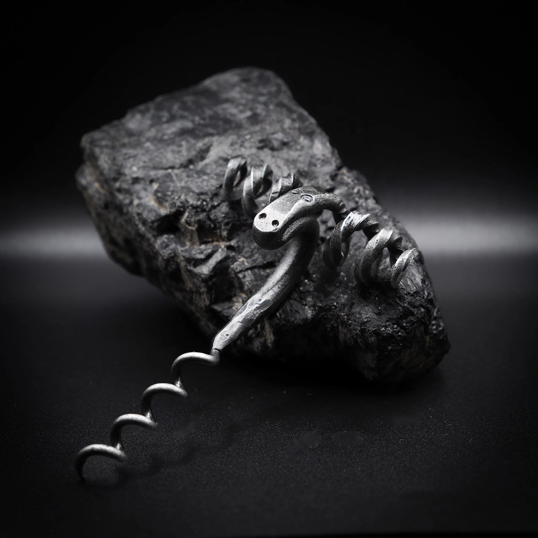 Handsmide bagge-skruv (råmskruv) på granitatsten. Gjord av återvunnet järn med mörkpatina och metalliska högdagrar. Baggehuvudet visar böjda horn som formar dramatisk handtag. Ansiktsfötryck och öga-detaljer synliga. Spiral skruv nedan. Dramatisk sidbelysning betonar metallartverk och hantverk.